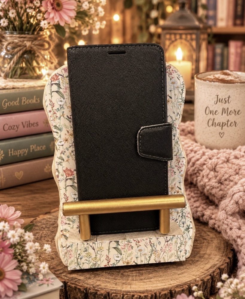 Handmade wooden phone holder with wavy edges, covered in a soft wildflower print, with a gold bar holding a phone upright beside books, a candle, and a Just One More Chapter mug